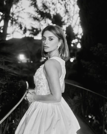 Penélope Cruz gazes over her shoulder in an eye-level medium shot, wearing an off-white gown with floral embellishments outdoors.