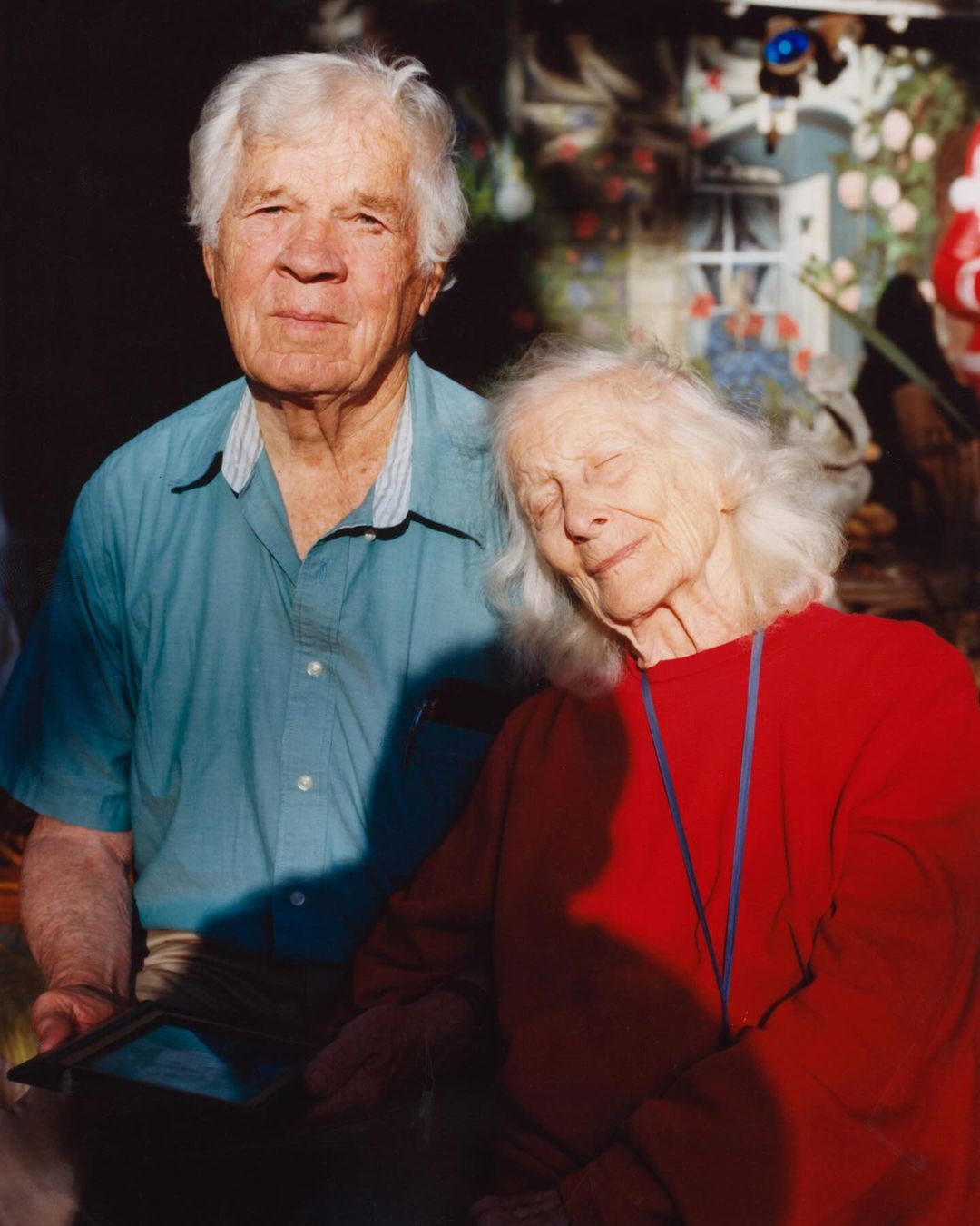 Portrait of an elderly couple in bright sunlight. The man with white hair wears a blue shirt and the woman a red top, with decorative background.