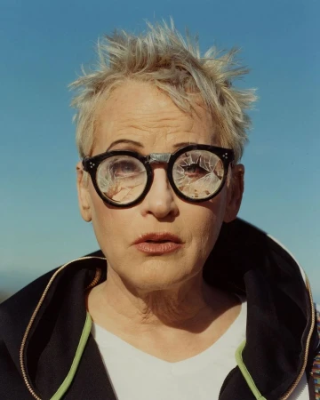 Portrait of woman with short blond hair wearing broken glasses against a bright blue sky background, wearing a white t-shirt and black jacket.