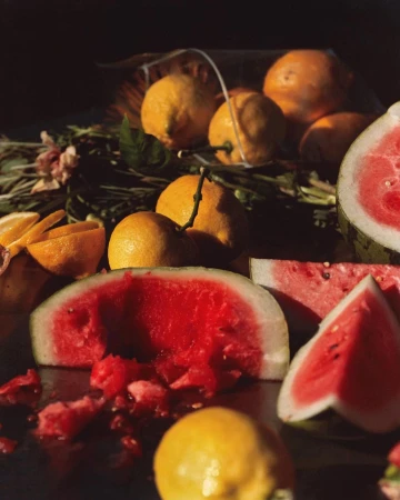 Vibrant still life featuring sliced watermelon, whole lemons, and oranges arranged with scattered blossoms in a naturally lit setting.