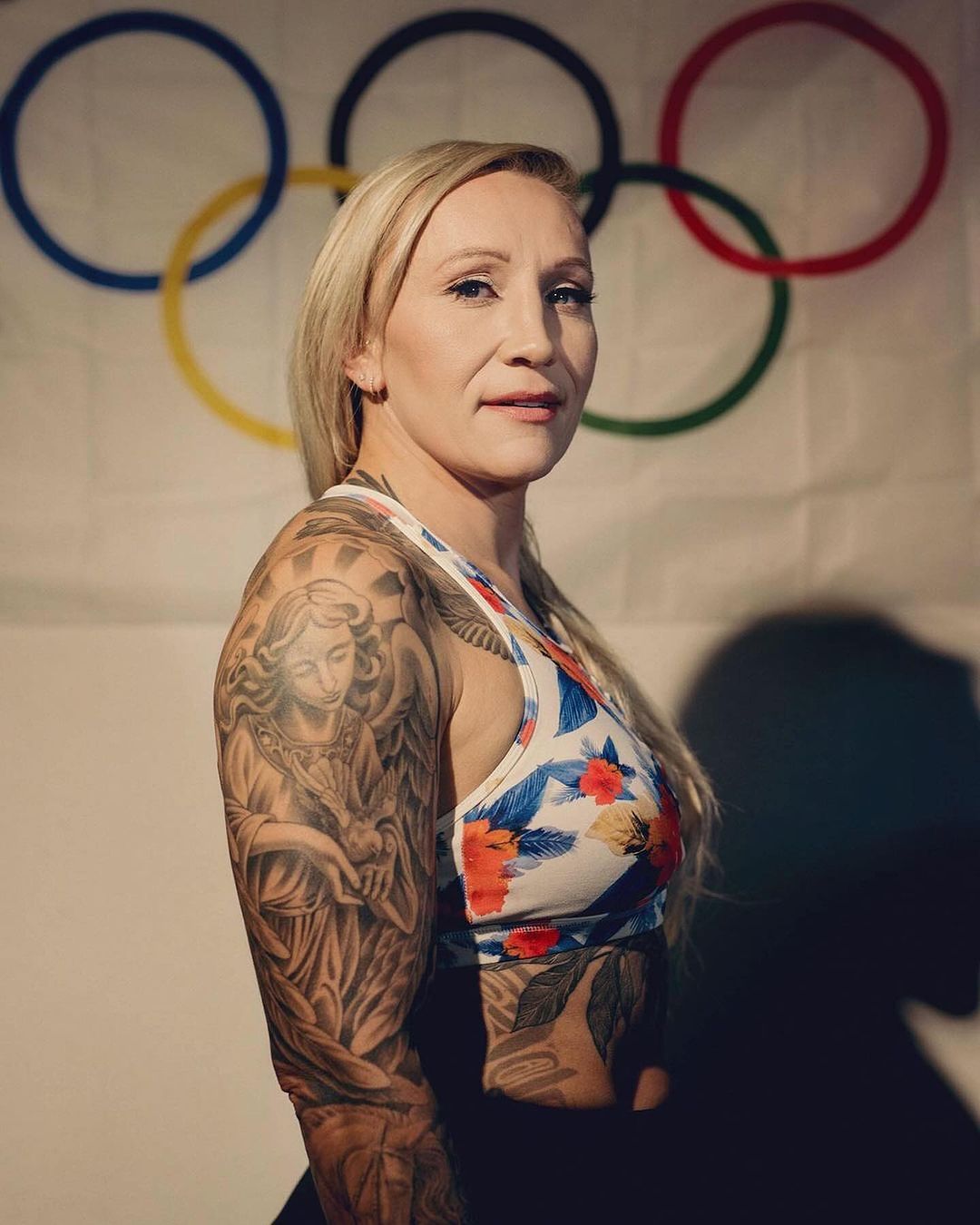 Blonde woman with angel tattoo posing in a floral top with Olympic rings in the background. Fitness and healthy lifestyle theme.