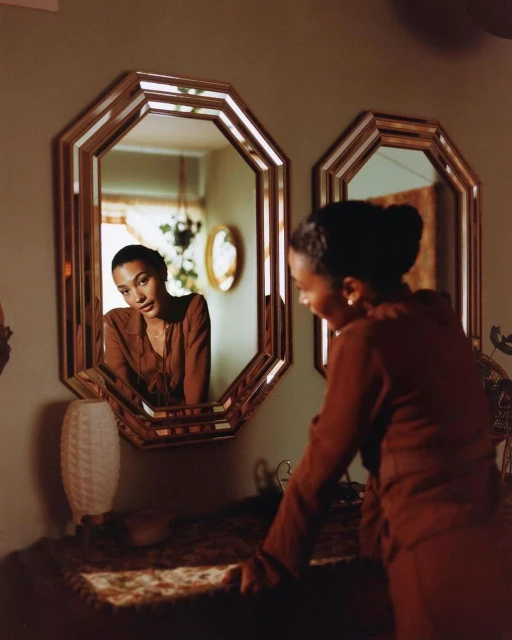 A woman in a brown outfit stands near a vanity with a mirror reflecting her face; the interior features warm tones and natural light.