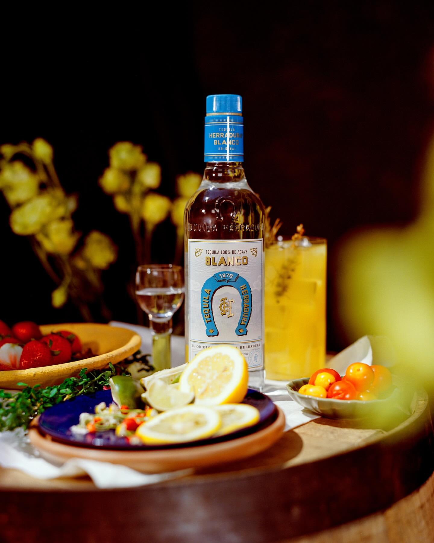 Bottle of Herradura Blanco Tequila surrounded by cocktail, shot glass, lemon wedges, and tomatoes on a wood barrel surface.