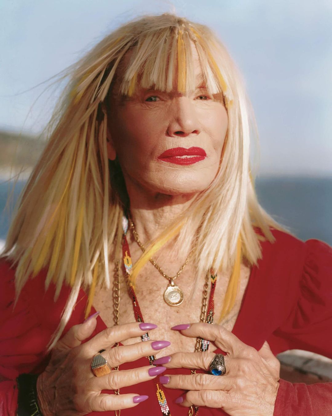 Elderly adult woman with blonde hair and red lipstick posing outdoors with layered necklaces and multiple rings, wearing a red top.