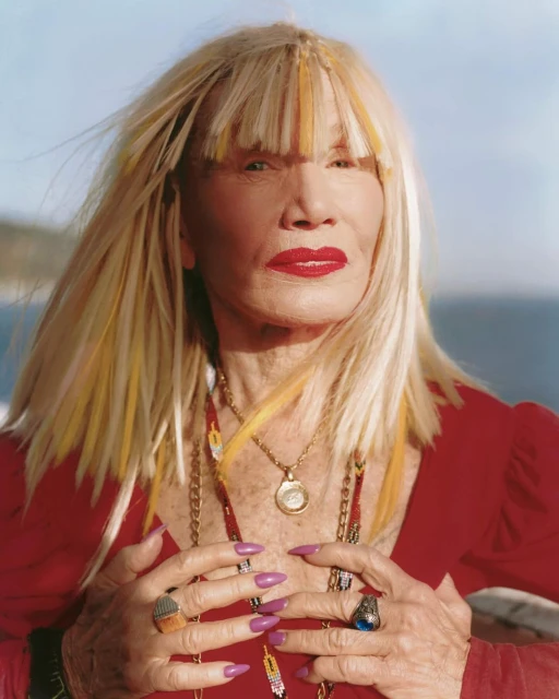Elderly adult woman with blonde hair and red lipstick posing outdoors with layered necklaces and multiple rings, wearing a red top.