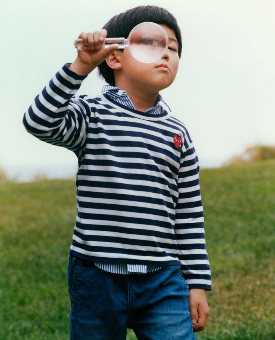 A young boy with dark hair stands outdoors on a grassy hill, holding a magnifying glass up to his eye in a medium, eye-level shot.