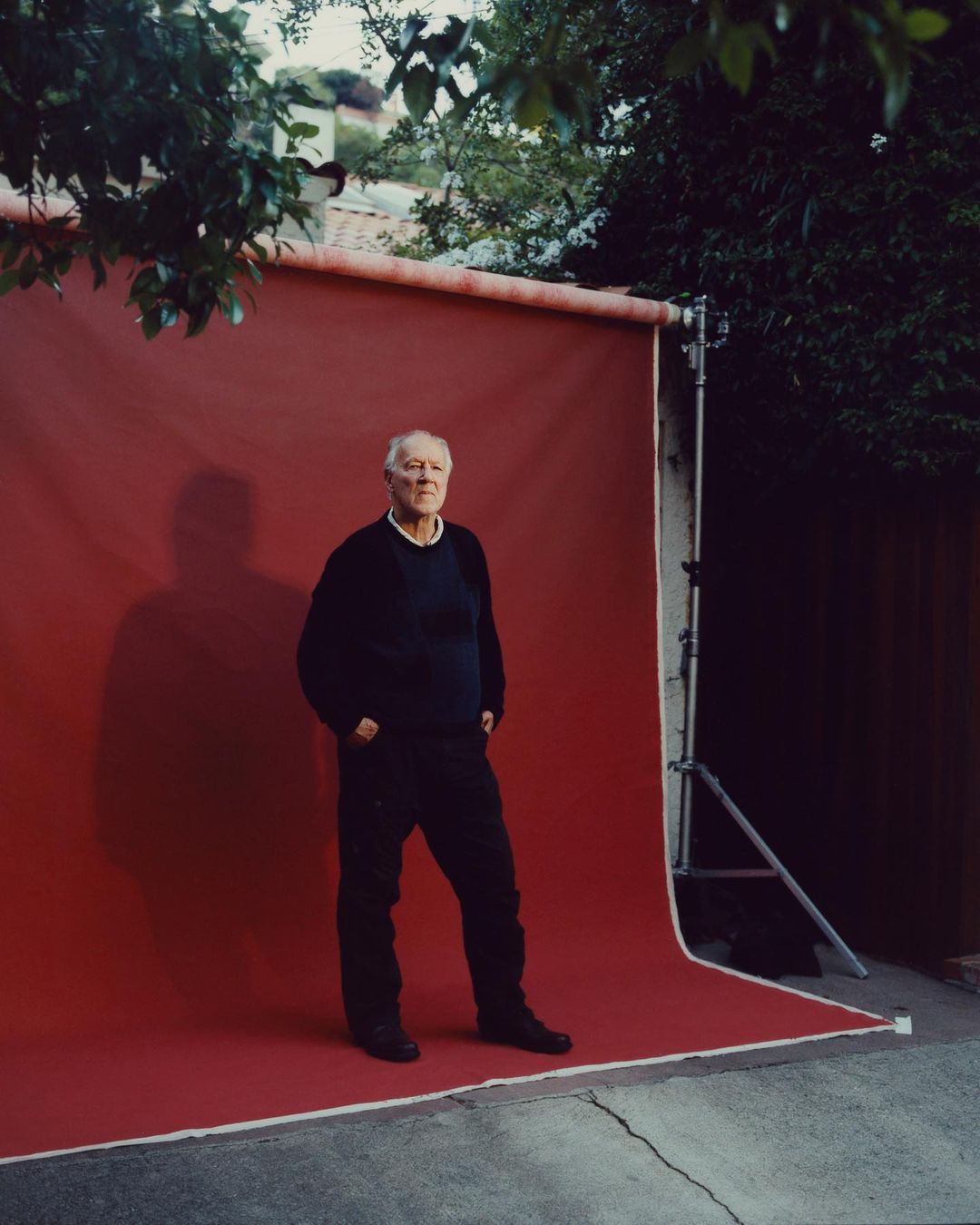 An elderly man in a black outfit poses confidently against a red backdrop in an outdoor setting, with lush foliage framing the scene.