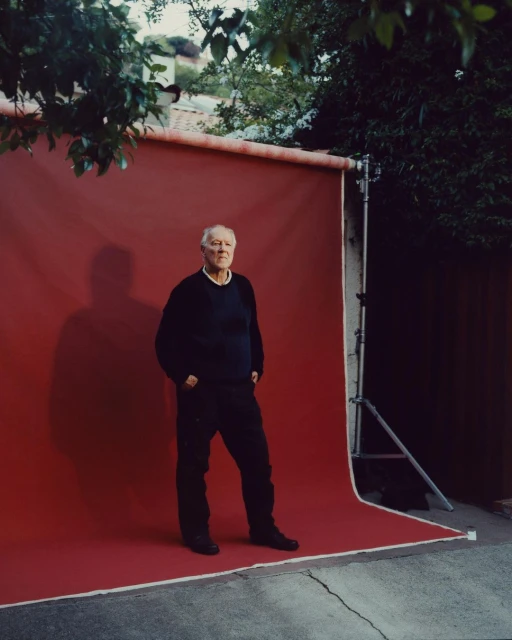 An elderly man in a black outfit poses confidently against a red backdrop in an outdoor setting, with lush foliage framing the scene.