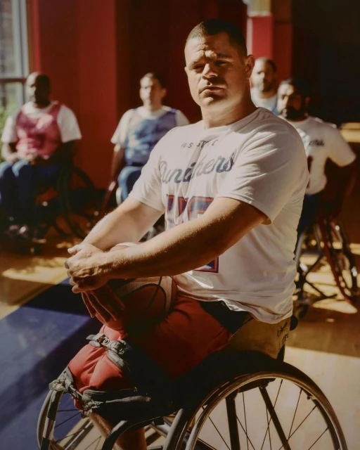Man in wheelchair with basketball and team athletes in background on the court. Indoor sports photography shows active and accessible lifestyle.