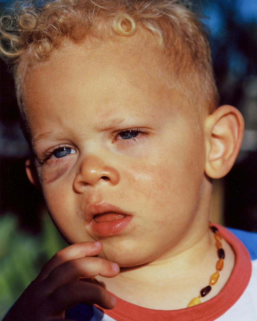 Close-up of a young child with blond curly hair, blue eyes, and a beaded necklace, gazing with a slightly concerned expression outdoors.