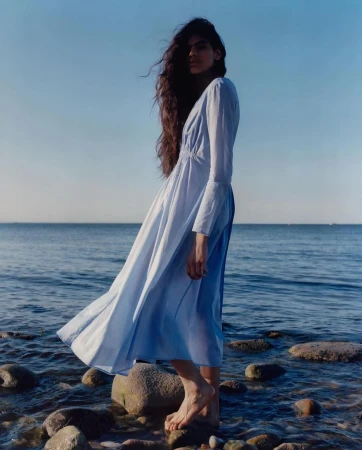 Woman with long dark hair wearing a light blue dress standing on rocky shore by the ocean