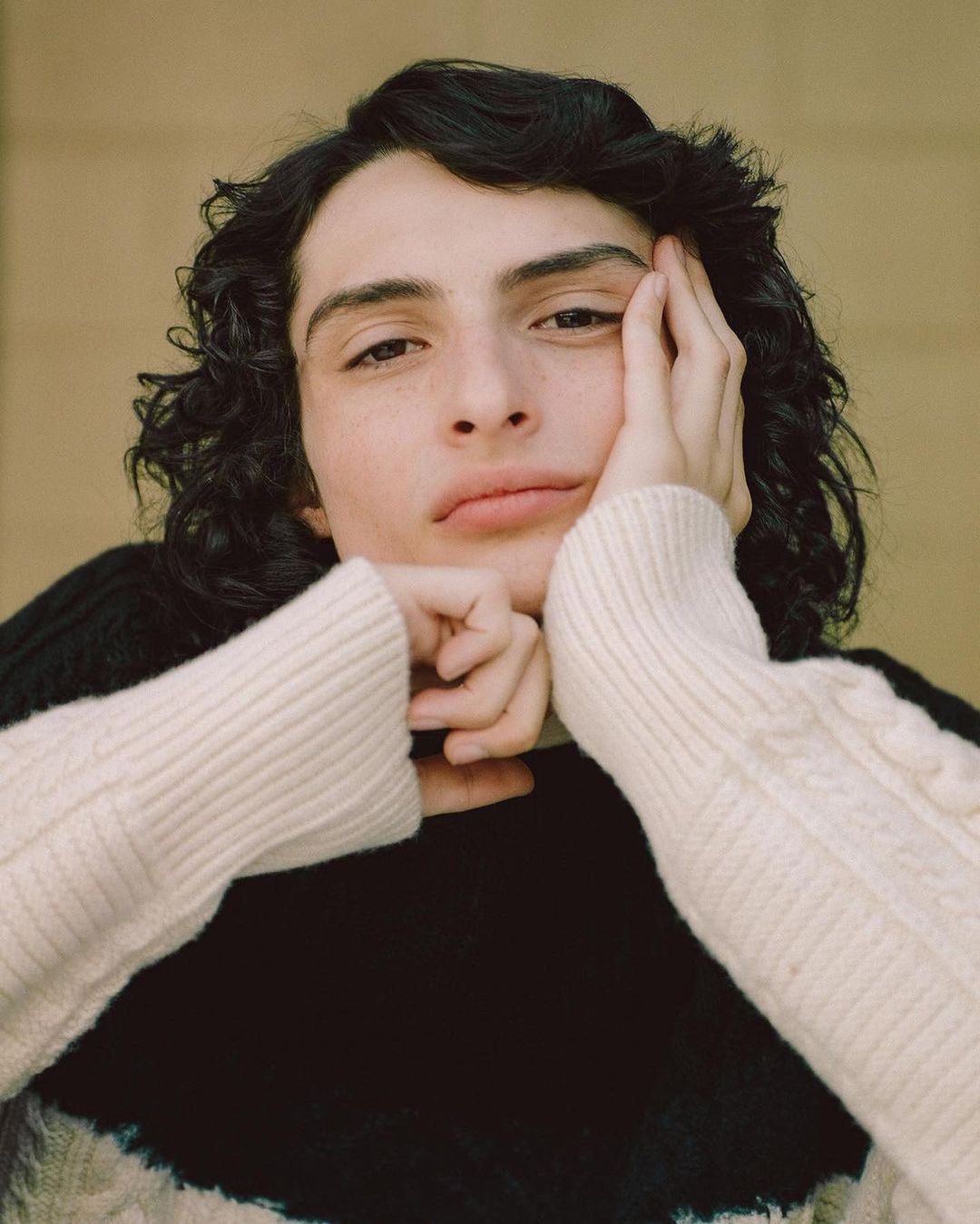 Close-up portrait of a young adult male with dark curly hair and a pensive expression, wearing a cream and black cable knit sweater.