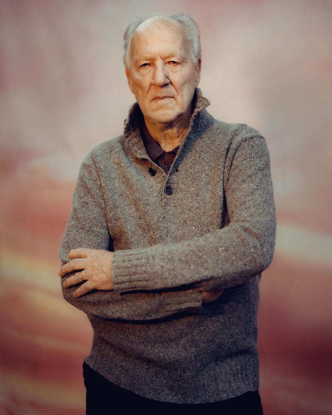 Portrait of a senior man in a grey sweater with arms crossed against a mottled pink backdrop.  He has short white hair and a serious expression.