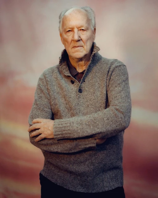 Portrait of a senior man in a grey sweater with arms crossed against a mottled pink backdrop.  He has short white hair and a serious expression.
