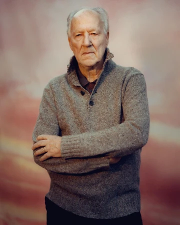 Portrait of a senior man in a grey sweater with arms crossed against a mottled pink backdrop.  He has short white hair and a serious expression.