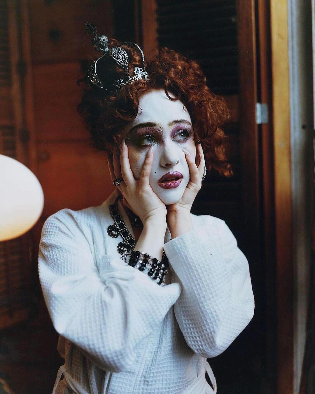 Portrait of a woman with dramatic makeup, a crown, and a white robe, holding her face with her hands, in an indoor setting.