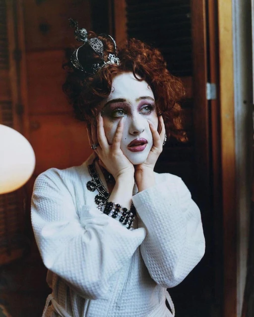 Portrait of a woman with dramatic makeup, a crown, and a white robe, holding her face with her hands, in an indoor setting.