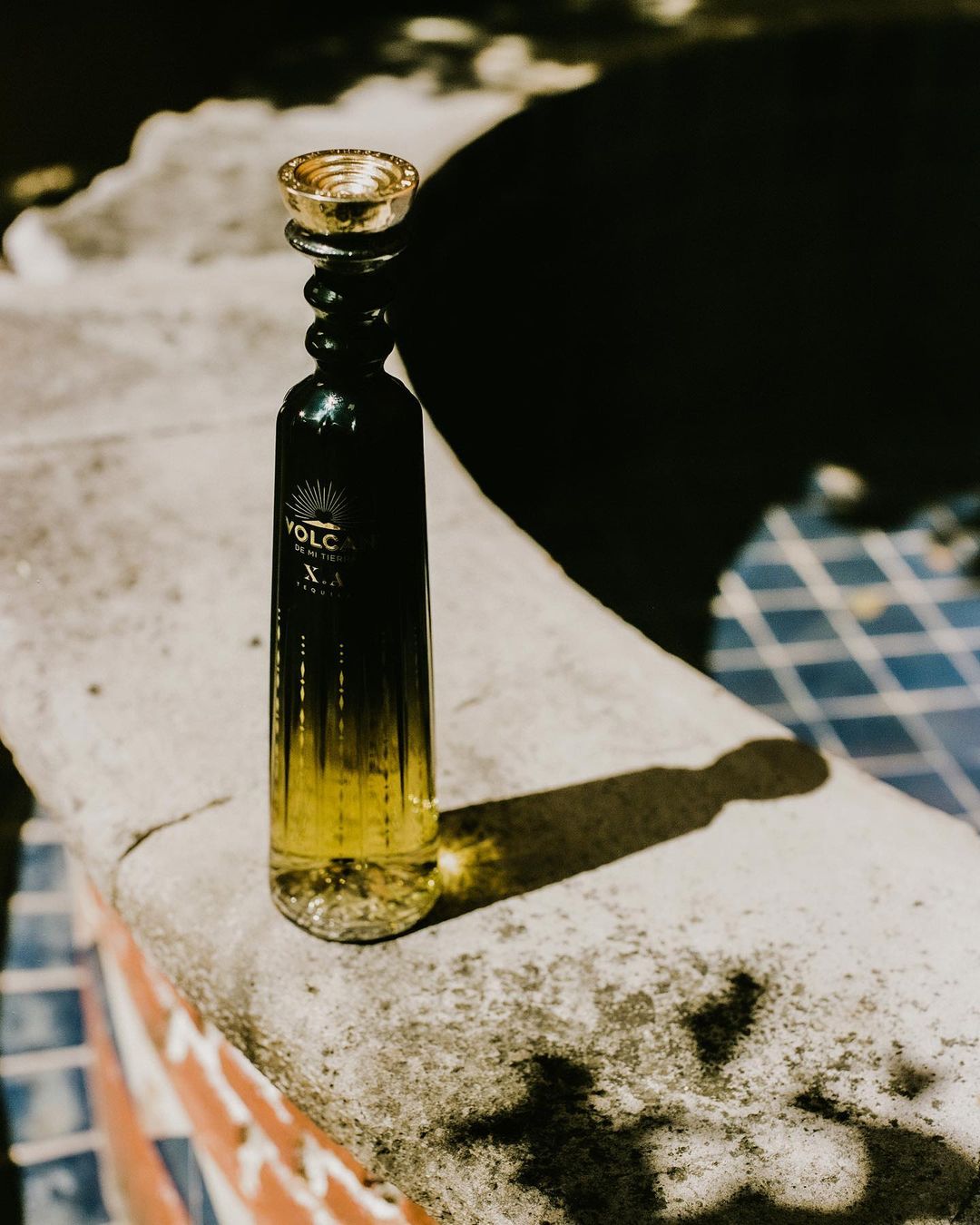 A bottle of Volcan X.A. tequila sits on a stone ledge in the sunlight, casting a shadow on the surface.