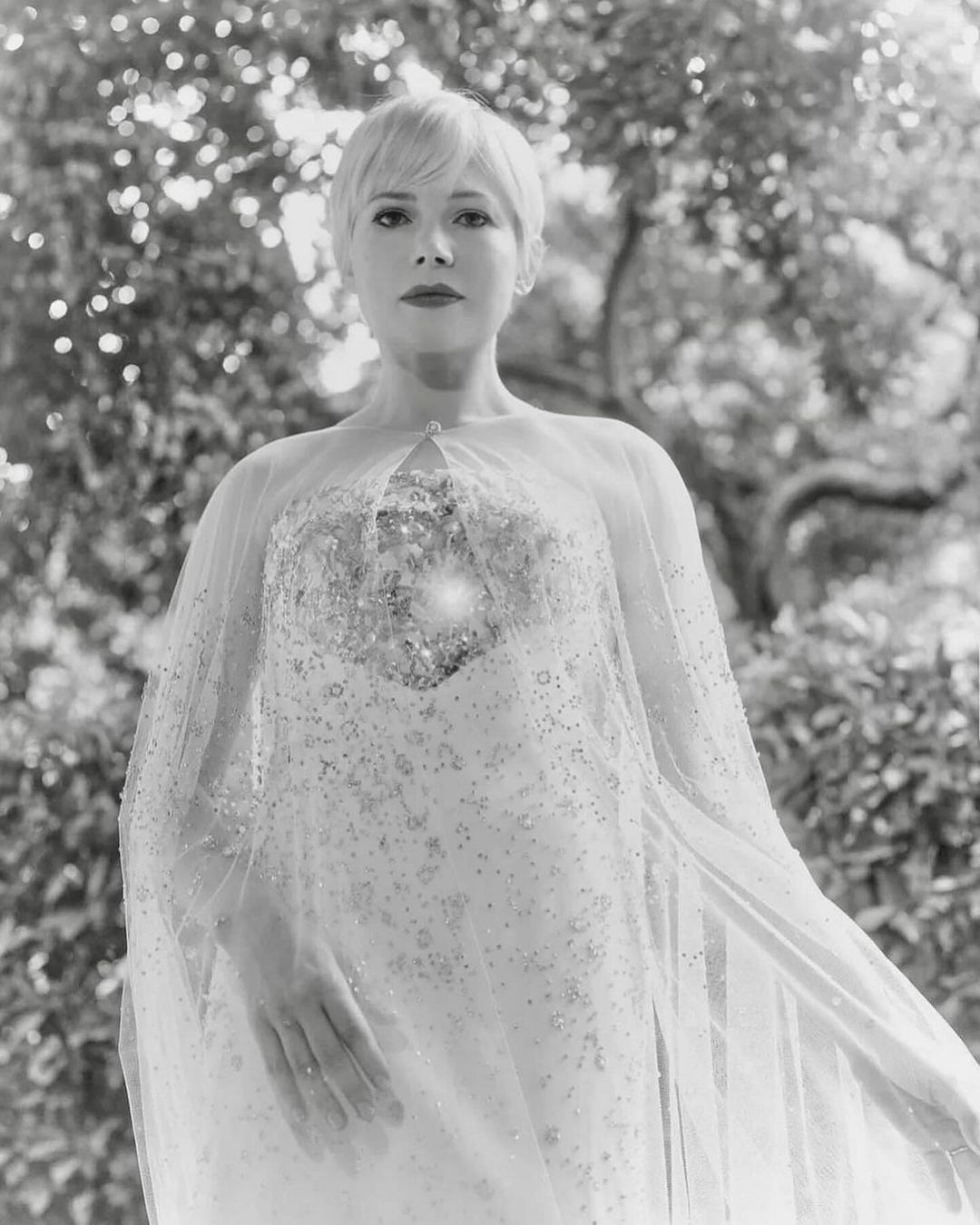 Monochrome portrait of a short-haired woman in an embellished gown and sheer cape, standing against a natural green foliage background.