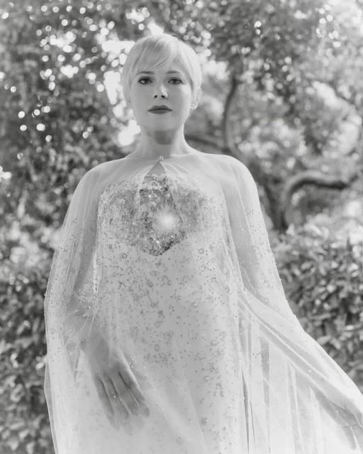 Monochrome portrait of a short-haired woman in an embellished gown and sheer cape, standing against a natural green foliage background.