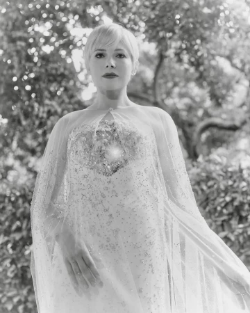Monochrome portrait of a short-haired woman in an embellished gown and sheer cape, standing against a natural green foliage background.