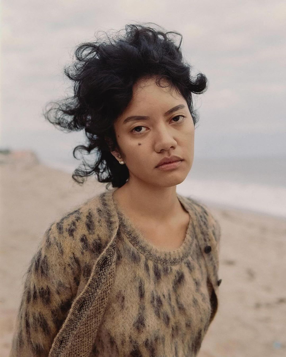 Portrait of a woman with dark curly hair wearing a leopard print cardigan on a sandy beach