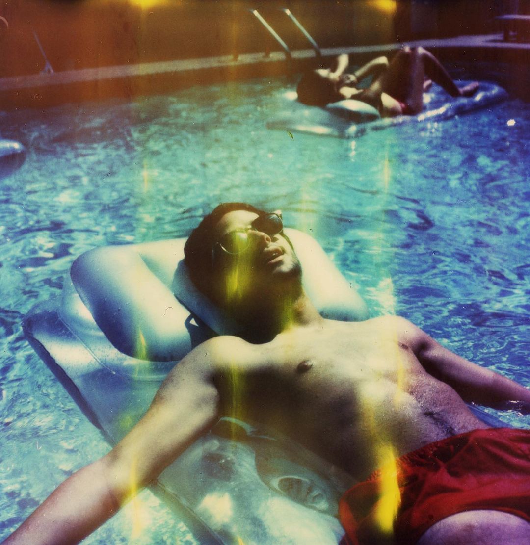 Man relaxes on an inflatable raft in a pool wearing sunglasses, while another person relaxes in the background on a raft as well.