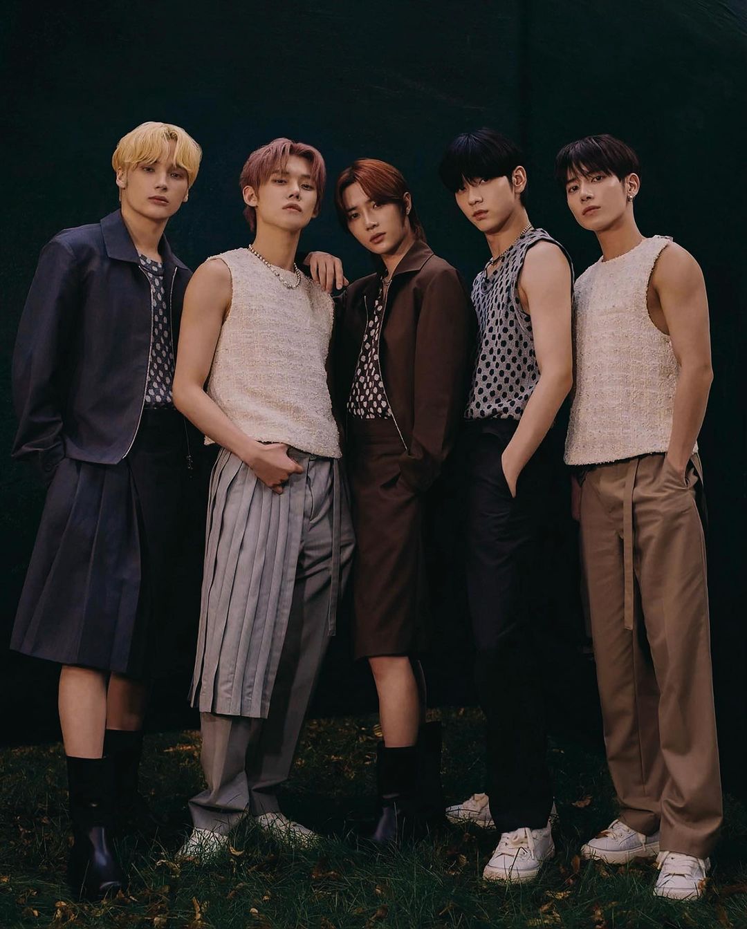 Group of five young adult men posing indoors in fashionable outfits, including pleated skirts, pants, and sleeveless tops against a dark backdrop.