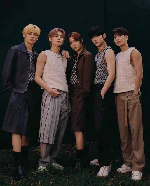 Group of five young adult men posing indoors in fashionable outfits, including pleated skirts, pants, and sleeveless tops against a dark backdrop.