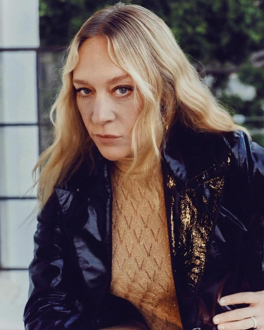 Portrait of Chloë Sevigny wearing a glossy black jacket and patterned beige top against a light background