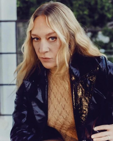 Portrait of Chloë Sevigny wearing a glossy black jacket and patterned beige top against a light background