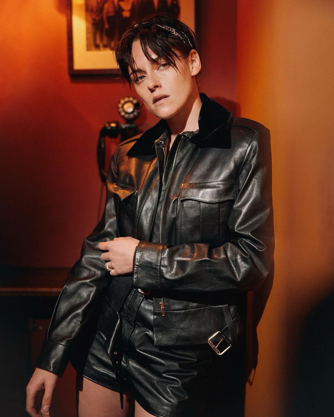 Portrait of an adult woman in a black leather jacket and skirt in an indoor setting with warm lighting, against a red and brown wall.