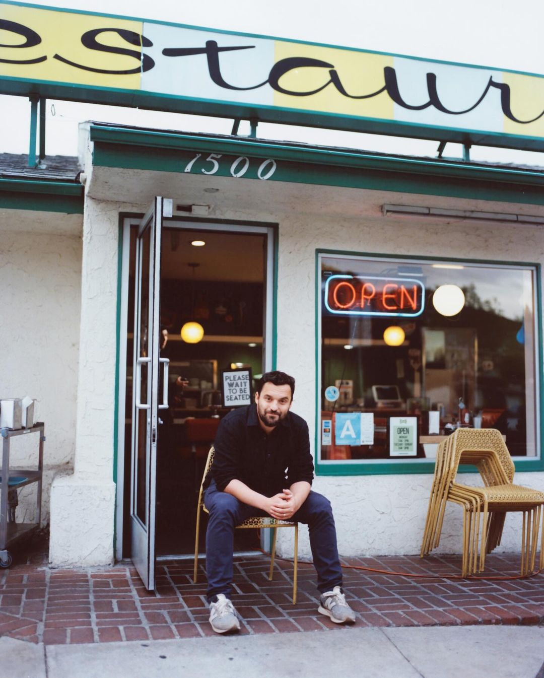 A man sits outside a restaurant in a black shirt and blue jeans on a wicker chair beside stacked chairs and an open sign.