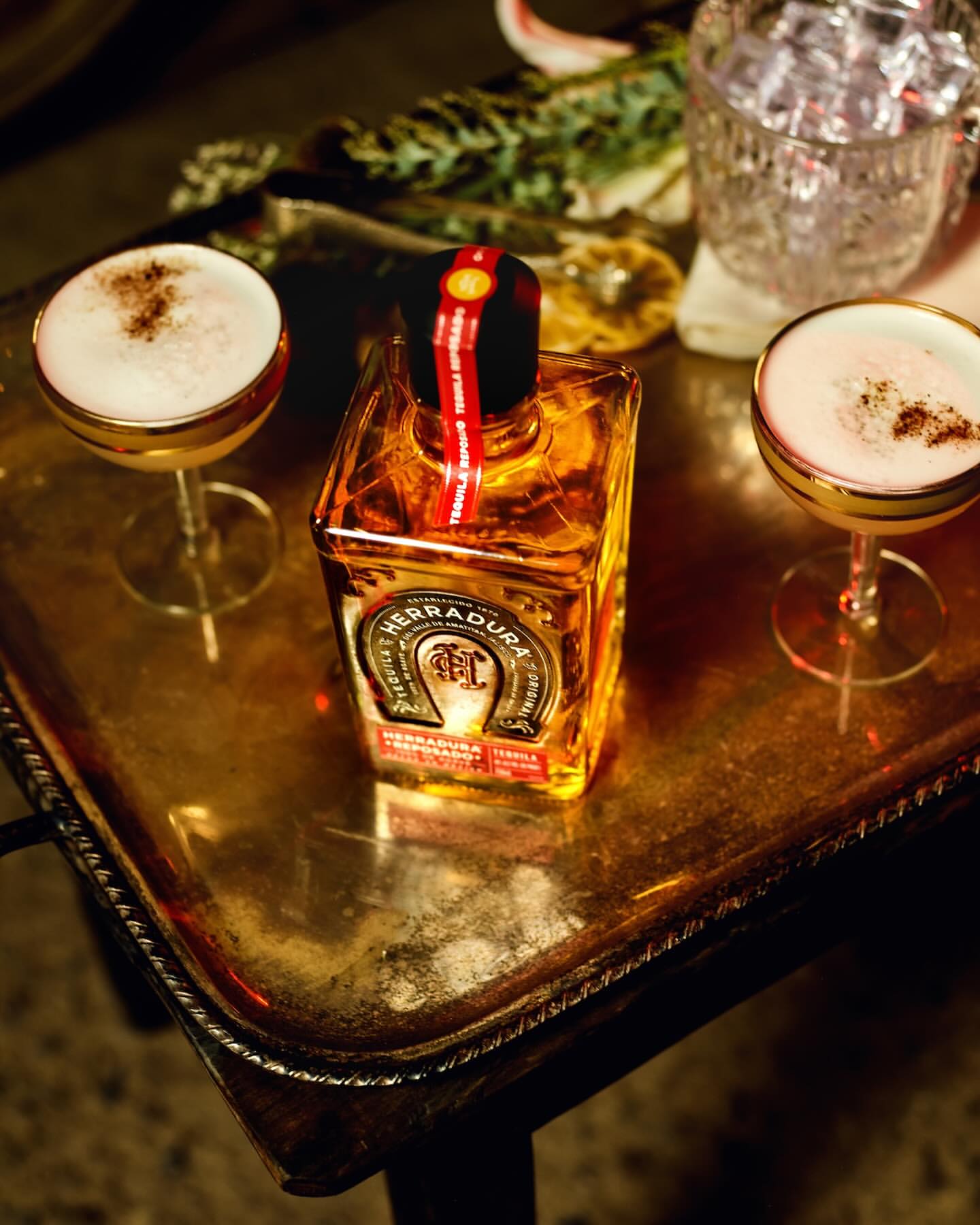 Tequila bottle and two stemmed cocktail glasses filled with yellow liquid and white foam sitting on an ornate silver tray.