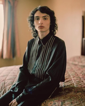 Portrait of an adult male with dark curly hair, wearing a black shirt with white stripes, sitting on a patterned bed in a room.