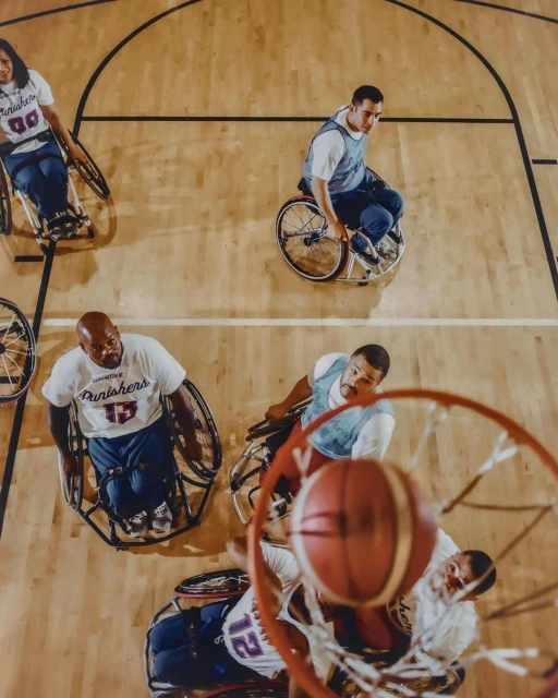 Overhead view of wheelchair basketball game in progress on hardwood court. Players in jerseys compete under hoop. Ball mid-air near net.