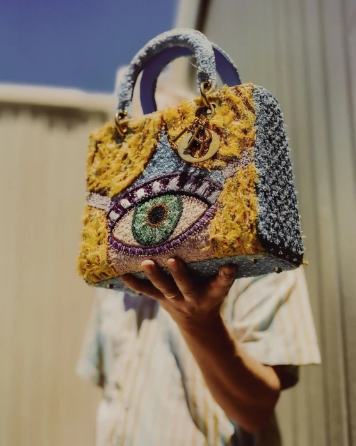 A close-up shot shows a person's hand holding an embroidered Dior handbag with an eye design against a sunny, outdoor backdrop.