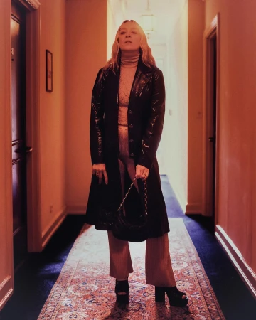 Woman standing in a hotel hallway wearing a dark leather trench coat, trousers, and platform sandals, holding a handbag.