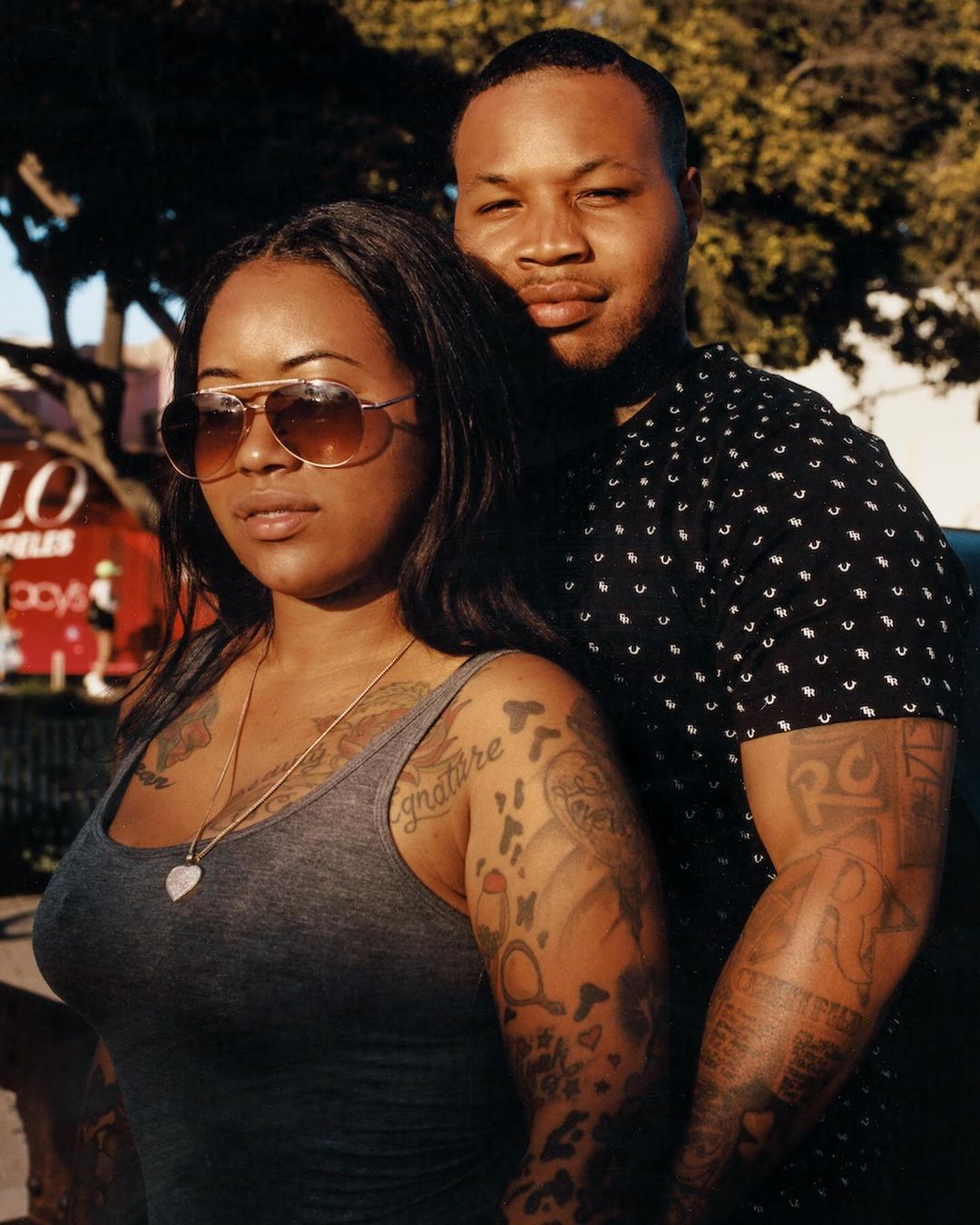 Portrait of a tattooed couple in urban setting, featuring a woman in sunglasses and gray tank top and a man in a patterned shirt.