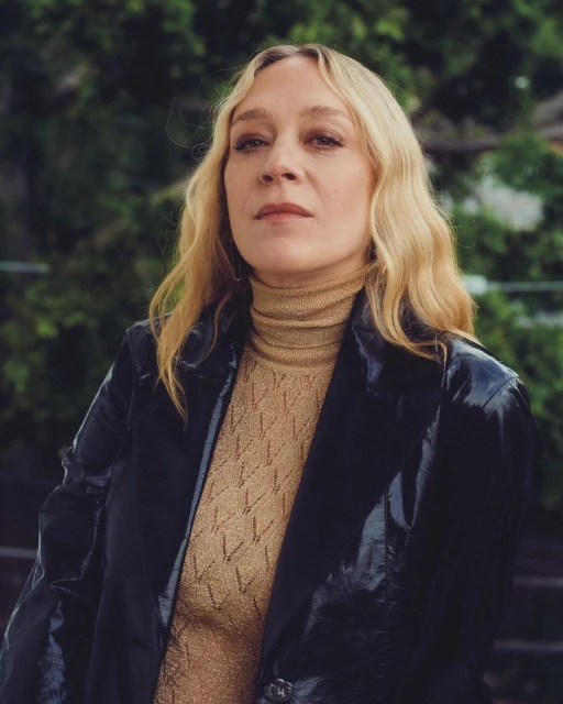 Close-up portrait of a blonde woman with fair skin wearing a patterned top and a black jacket against an outdoor green background.
