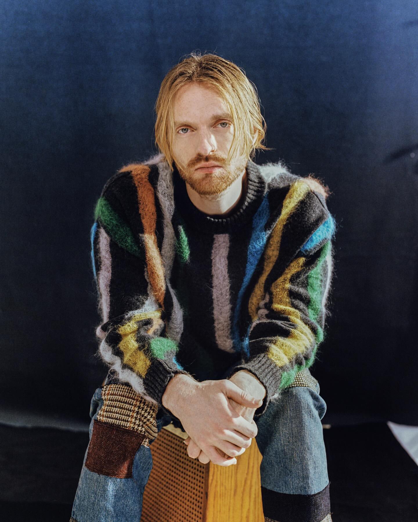 Man with blonde hair wearing a multicolored striped fuzzy sweater seated on a wooden stool against a dark blue backdrop