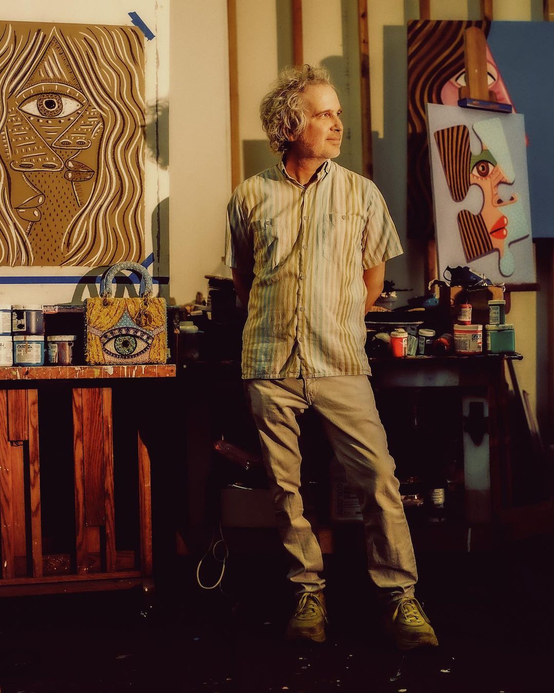 Artist with curly hair stands in his art studio, surrounded by painted canvases, art supplies, and a beaded handbag with an eye design.