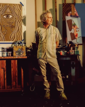 Artist with curly hair stands in his art studio, surrounded by painted canvases, art supplies, and a beaded handbag with an eye design.