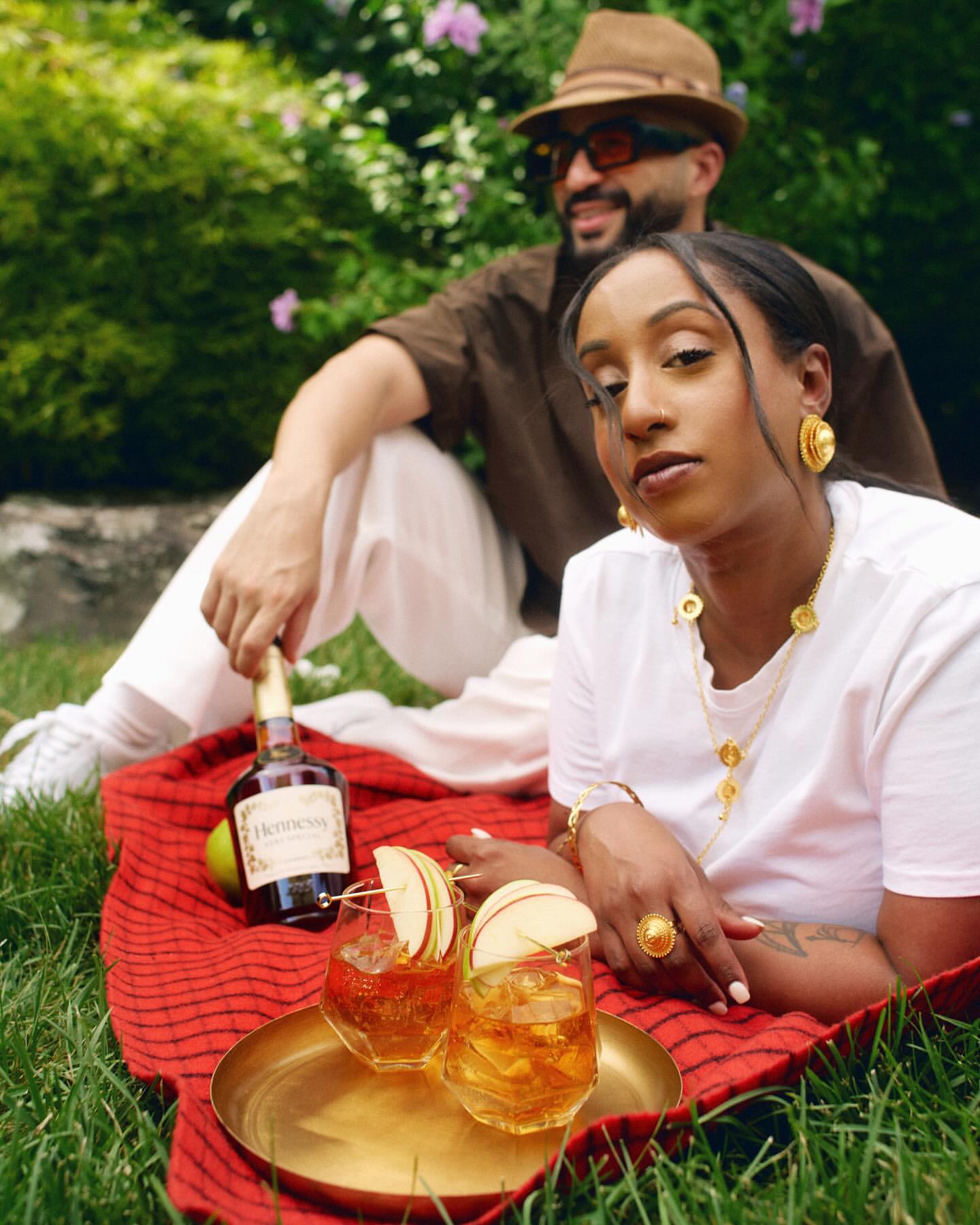 Woman and man enjoying Hennessy cocktails in the park, sitting on a red blanket, surrounded by greenery, wearing fashionable attire.