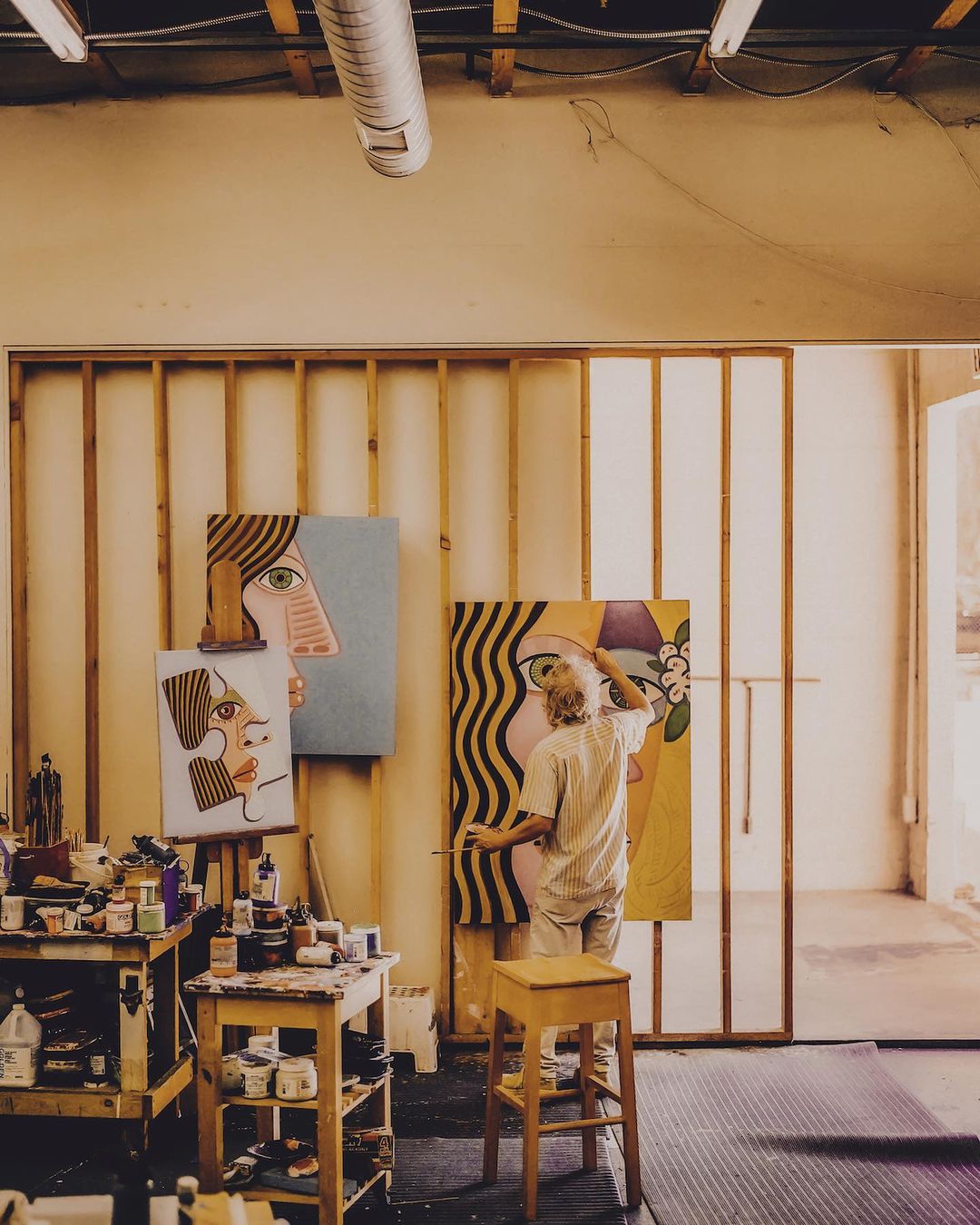 Artist in studio working on large portrait paintings with visible paint cans and wooden easel, bathed in soft natural light.