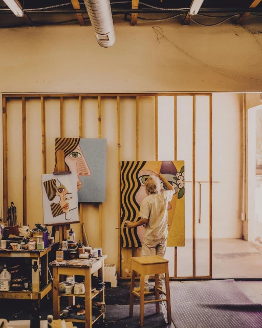 Artist in studio working on large portrait paintings with visible paint cans and wooden easel, bathed in soft natural light.