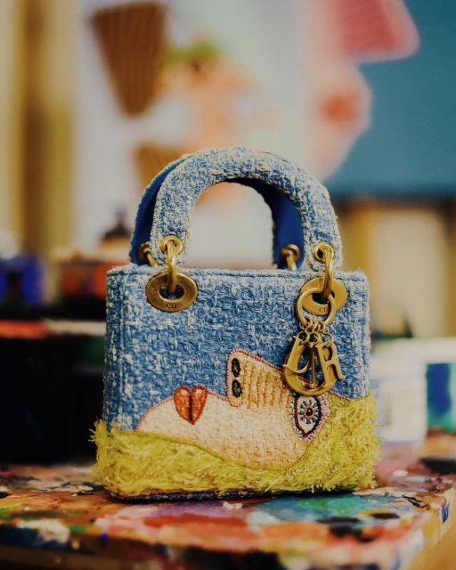 Close-up of a light blue tweed Lady Dior micro bag with gold hardware and an abstract design on an artistically-painted table.