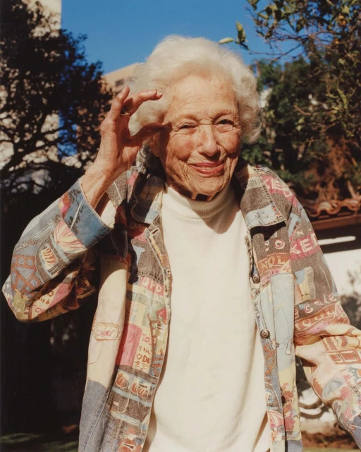 An elderly woman with white hair smiles and winks, holding her hand near her eye in an outdoor setting with trees and a building.