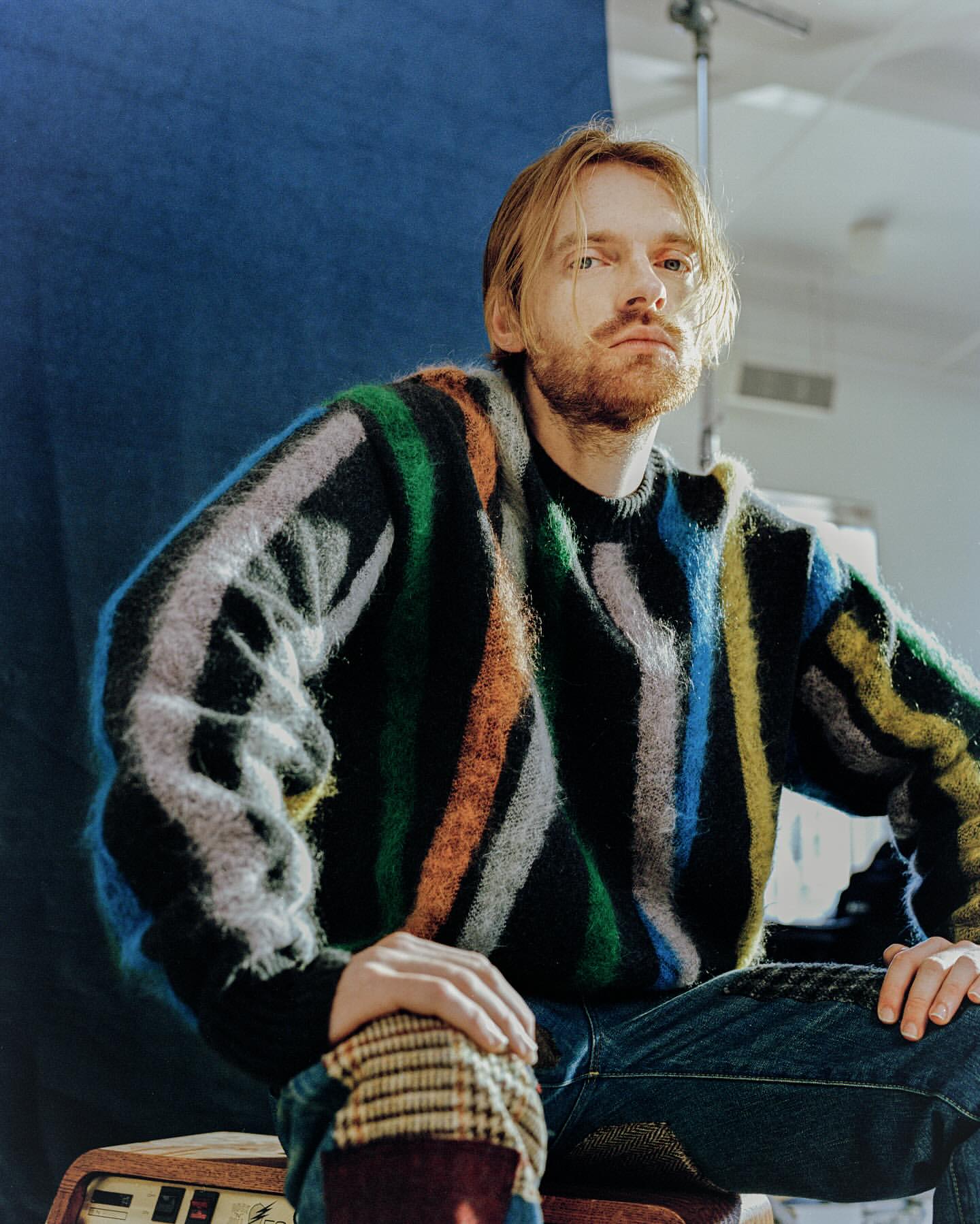 Portrait of a blond man with a beard wearing a colorful striped sweater sitting indoors against a blue and white wall.