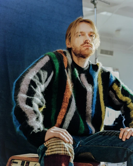 Portrait of a blond man with a beard wearing a colorful striped sweater sitting indoors against a blue and white wall.